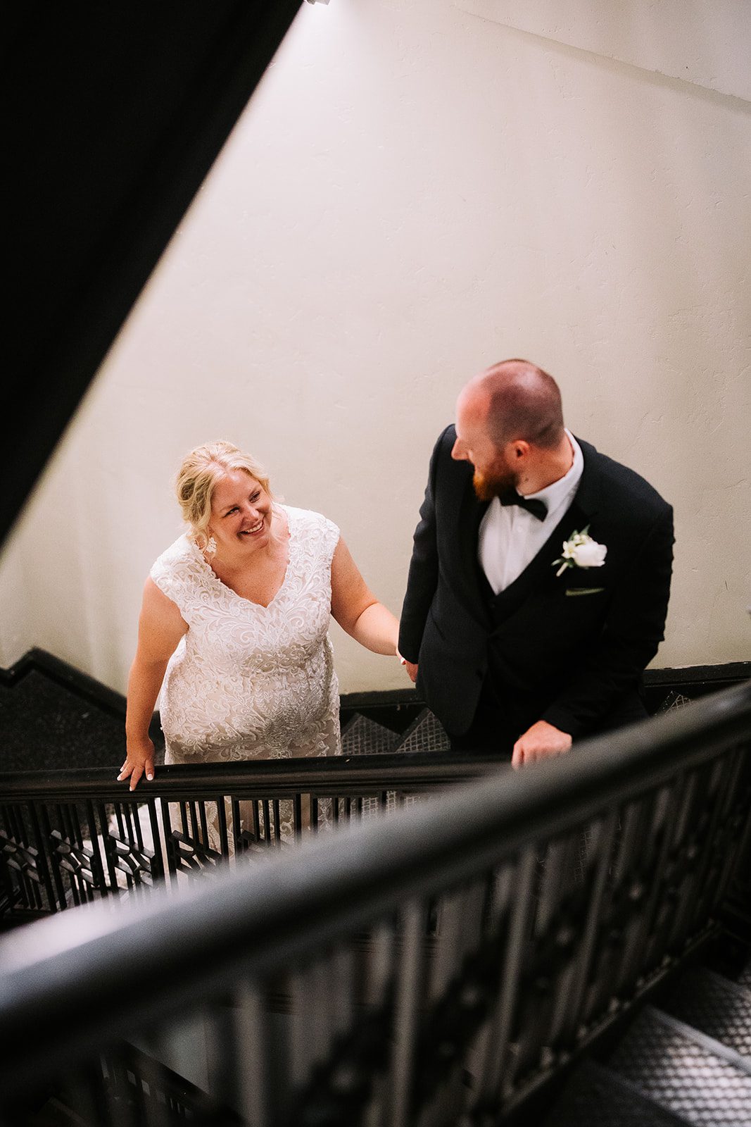 bride and groom walking up stairs with him leading her looking at eachother with loving eyes with editorial lighting in hallway