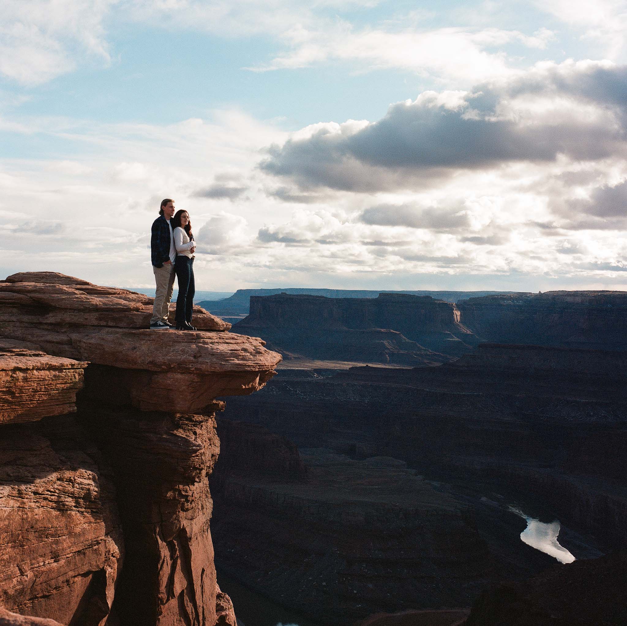 Couple standing on a narrow rock ledge overlooking a vast canyon under a partly cloudy sky